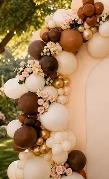Arco de globos orgánico en chocolate, crema y dorado con rosas, en un jardín al atardecer.