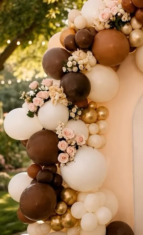 Arco de globos en chocolate, crema y dorado con rosas sobre un backdrop de arco, en un jardín.