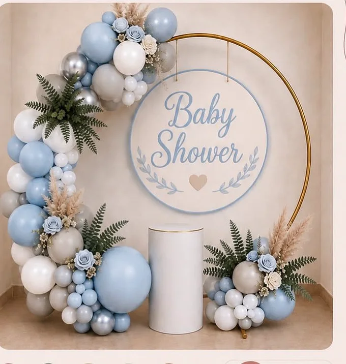 Aro metálico dorado con letrero «Baby Shower», guirnalda orgánica de globos en azul, gris y blanco, follaje y cilindro para el pastel.
