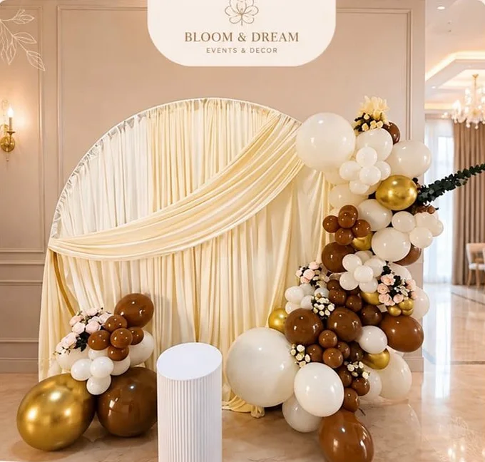 Backdrop redondo con tela drapeada en ivory y guirnalda orgánica de globos en blanco, caramelo, chocolate y dorado, con pedestal cilíndrico.